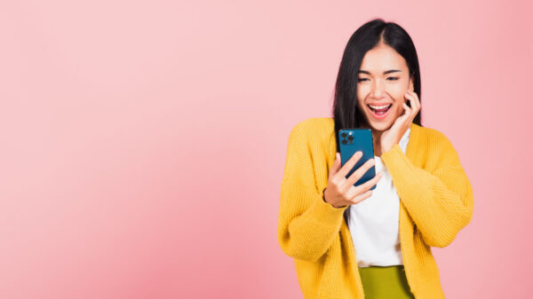 Retrato de uma mulher feliz, sorrindo, animada, surpresa, usando um celular, em um estúdio isolado em um fundo rosa.
