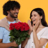 homem segurando um buque de rosas e entregando para uma mulher em fundo amarelo