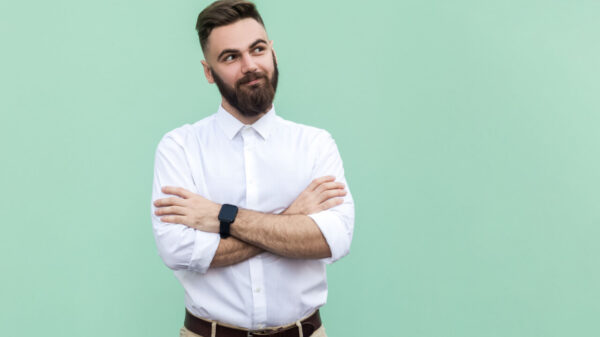 Retrato de um homem barbudo, usando uma camisa branca, em pé, com os braços cruzados, olhando para o lado, com um leve sorriso. Foto de um estúdio interno isolada em um fundo verde claro.