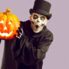 Homem usando fantasia de um esqueleto com uma capa preta e uma cartola para o Halloween. Isolado em um fundo roxo claro, segurando uma abóbora iluminada e olhando para a câmera com uma expressão surpresa no rosto.