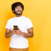 Homem carismático, de camiseta branca, segurando um celular nas mãos, olhando alegremente para o lado, pensando, em pé sobre um fundo laranja isolado, sorrindo.