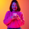 Mulher de suéter rosa usando o celular sobre um fundo laranja gradiente em luz neon. Conceito de emoções, expressão facial e inspiração.