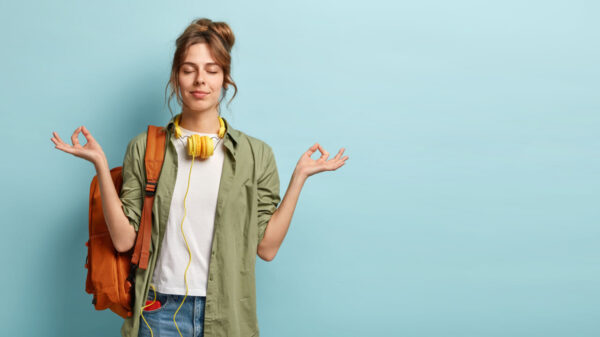 Foto de uma estudante relaxada, mantendo as mãos em um gesto de mudra, mantendo os olhos fechados, com fones de ouvido no pescoço, com uma mochila, isolada em um fundo azul.