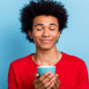 Retrato de um homem pacífico e positivo, com os olhos fechados, com as mãos segurando uma xícara de café, isolado em um fundo de cor azul.