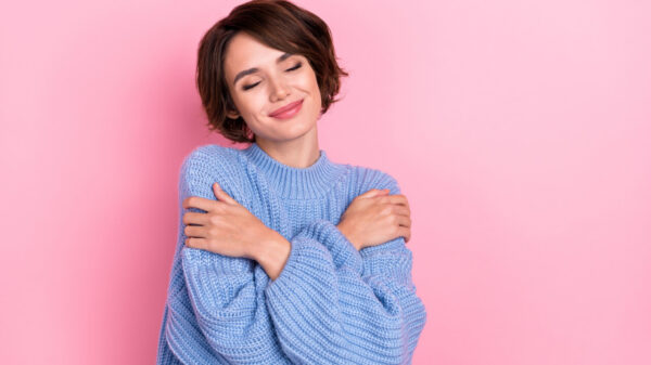 Retrato de uma mulher com os olhos fechados, abraçando os próprios ombros, isolada em um fundo de cor rosa.
