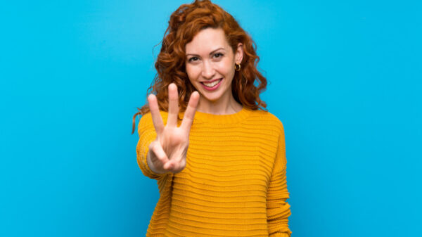 Mulher ruiva usando um suéter amarelo, feliz e fazendo o número três com os dedos.