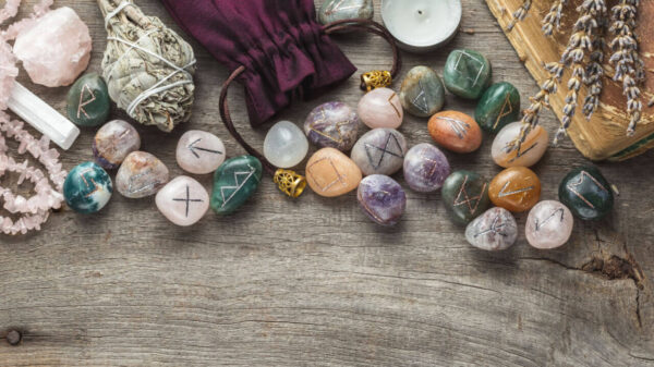 Conjunto de runas Elder Futhark feitas de pedras preciosas naturais, junto com um incenso de sálvia, uma vela e um livro antigo em um fundo de madeira.