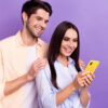 Foto de um homem e uma mulher positivos, juntos, olhando para a tela de um celular, isolados em um fundo de cor violeta.