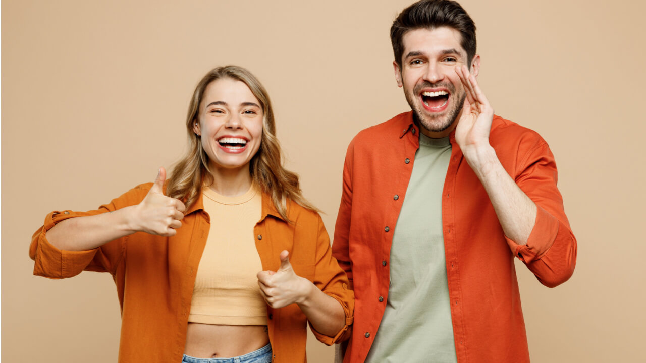 Casal composto por um homem e uma mulher usando roupas casuais. O homem gritando, compartilhando notícias, com uma mão perto da boca. Mulher com os polegares para cima. Isolados em um fundo de cor bege claro liso.