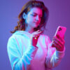 Mulher usando celular, lendo algo, contra um fundo de estúdio roxo e rosa em luz neon.