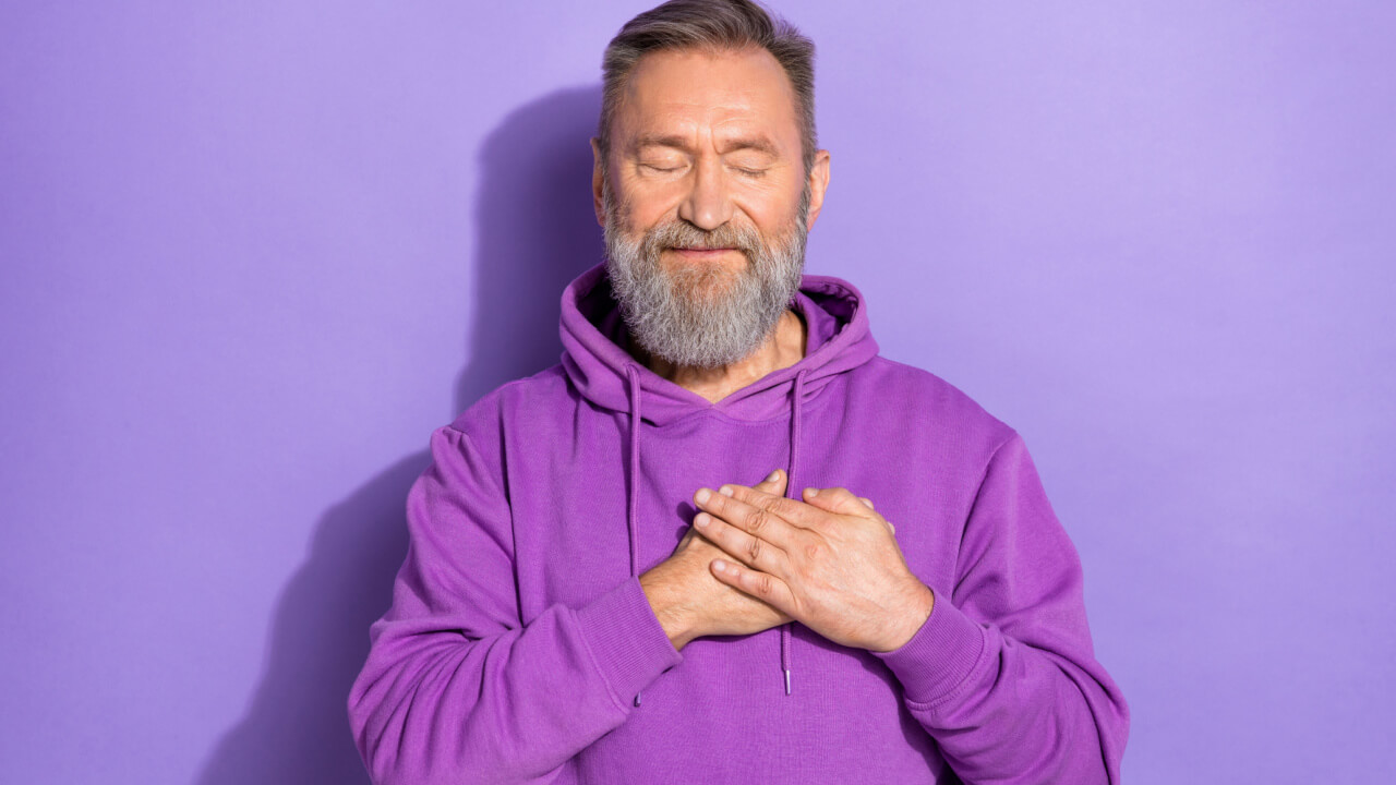 Foto de um homem usando um casaco com capuz casual, de olhos fechados, com as mãos sobre o coração, isolado em um fundo de cor roxa pastel.