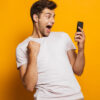 Retrato de um homem animado olhando para o celular, isolado sobre um fundo amarelo, celebrando.