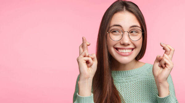 mulher sorrindo e cruzando os dedos em fundo rosa