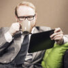 Homem sentado em um sofá, tomando um café e olhando para a tela de um tablet.