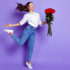 Foto de uma mulher na ponta do pé, segurando um buquê de rosas, com a boca aberta, usando uma camisa branca, em um fundo de cor violeta isolado.