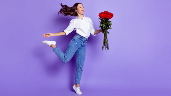 Foto de uma mulher na ponta do pé, segurando um buquê de rosas, com a boca aberta, usando uma camisa branca, em um fundo de cor violeta isolado.
