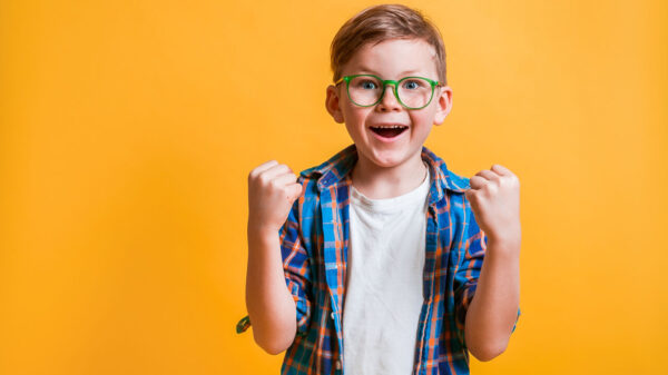 Menino usando óculos, isolado em um fundo amarelo. Garoto muito feliz e animado, fazendo gesto de vencedor com os braços levantados, sorrindo e gritando. Conceito de celebração.