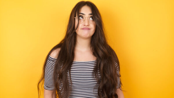 Mulher com cabelos longos, vestindo uma camiseta listrada, olhando para cima, com os lábios juntos, chateada, pensando em algo importante, com os braços para baixo.