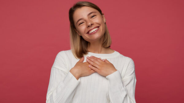 Retrato de estúdio de uma mulher posando sobre um fundo vermelho, com a mão no coração, sorrindo amplamente e se sentindo feliz.