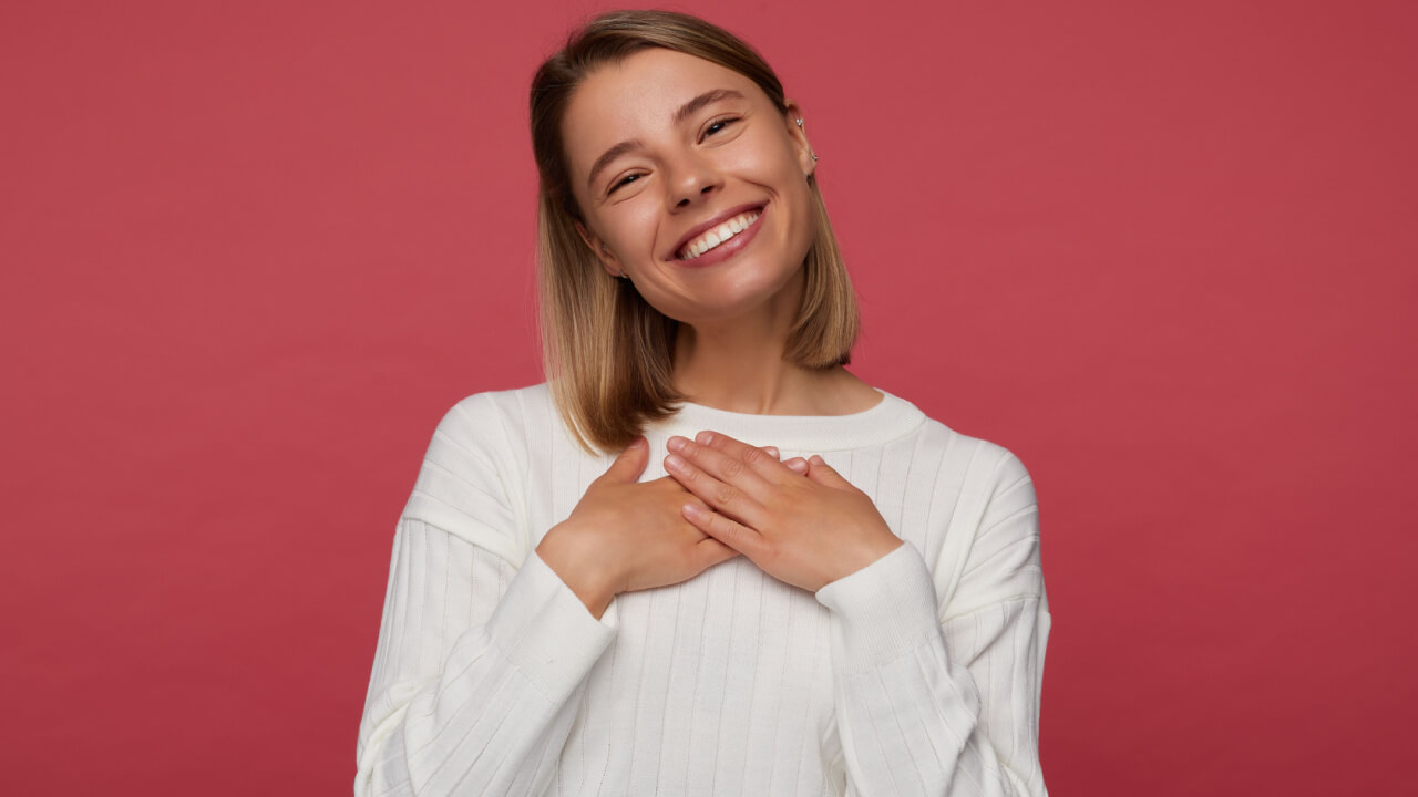 Retrato de estúdio de uma mulher posando sobre um fundo vermelho, com a mão no coração, sorrindo amplamente e se sentindo feliz.