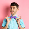 Homem fixando uma gravata borboleta no pescoço e sorrindo, sobre um fundo rosa.