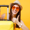 Mulher turista usando roupas casuais de verão, com um chapéu e uma mala, mostrando polegar para cima, isolada em um fundo amarelo. Conceito de viagem de voo aéreo.