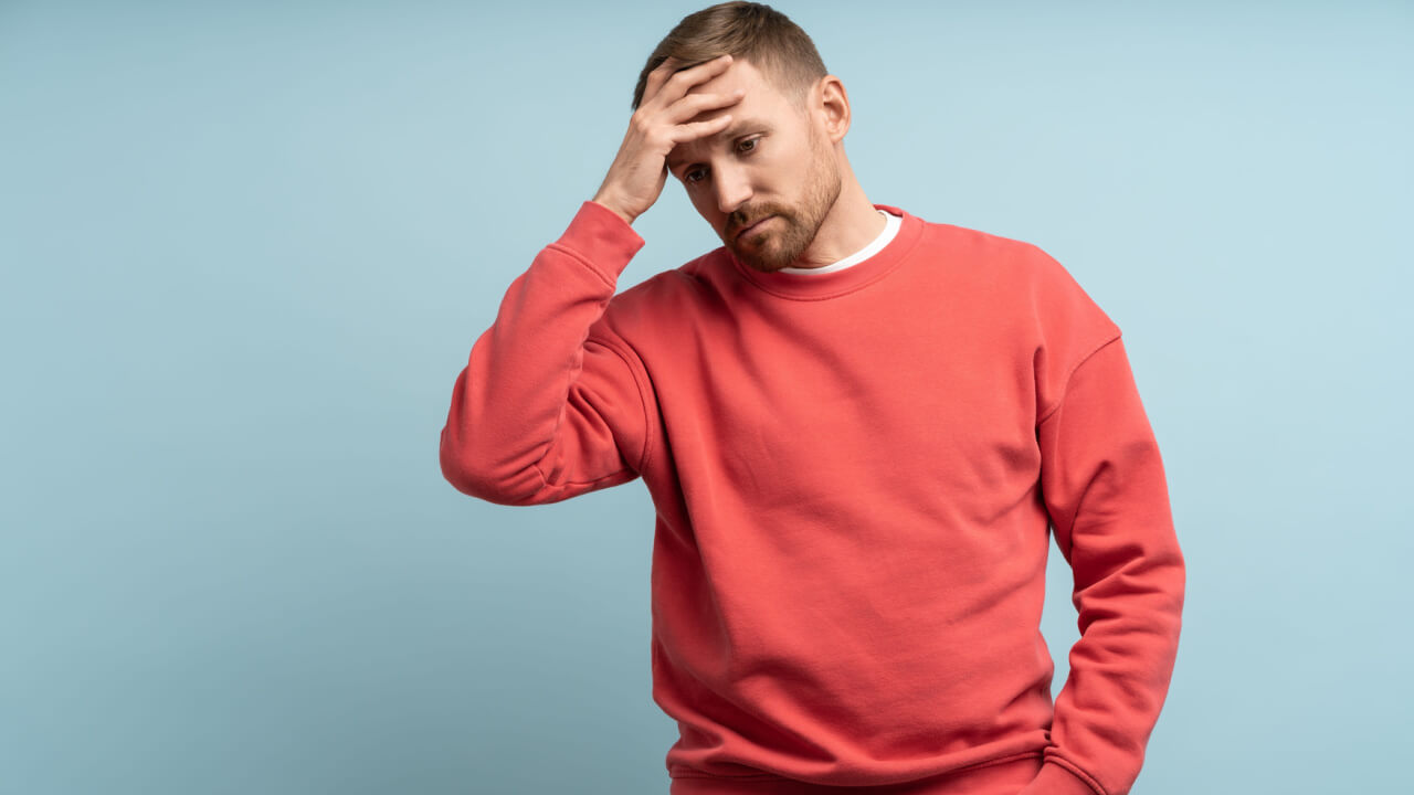 Homem preocupado, com a mão na testa, isolado em um fundo azul de um estúdio com uma expressão de decepção no rosto.