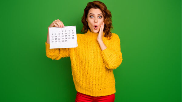 Foto de uma mulher muito chocada, segurando um calendário de papel, mostrando-o, com a mão na bochecha, usando um suéter de malha amarelo e calças vermelhas, isolada em um fundo de cor verde.