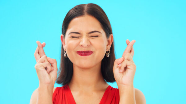Mulher feliz, com os dedos cruzados, sobre um fundo azul de um estúdio, desejando boa sorte, entusiasmada, otimista.