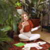 mulher sentada no chão segurando um livro em um ambiente cheio de plantas