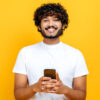Homem positivo, usando um celular, navegando na Internet, parado em um fundo amarelo isolado, olhando para a câmera, sorrindo.