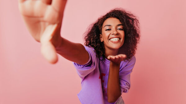 mulher negra brincando com a camera vestindo roxo e em fundo rosa