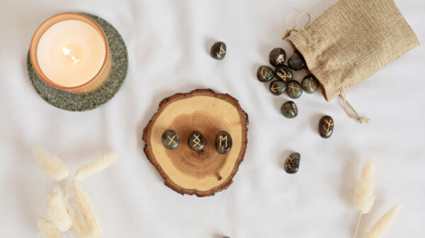 em cima de um lençol branco está uma madeira com uma vela e uma madeira com pedras rúnicas e também há um saquinho com pedras rúnicas
