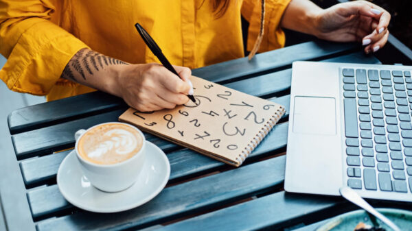 mulher sentada apoiada numa mesa de madeira com um bloco escrevendo números o notebook e uma xícara de café
