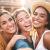 três amigas sorrindo tirando foto a céu aberto