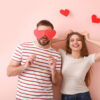 Casal feliz, segurando palitos com um papel vermelho em formato de coração na ponta, em um fundo rosa.