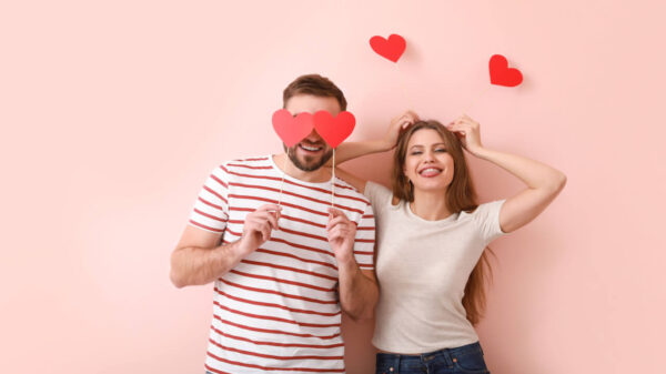Casal feliz, segurando palitos com um papel vermelho em formato de coração na ponta, em um fundo rosa.