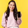 mulher de cabelo preto um pouco acima da cintura e segurando um celular com a tela virada para frente em fundo amarelo