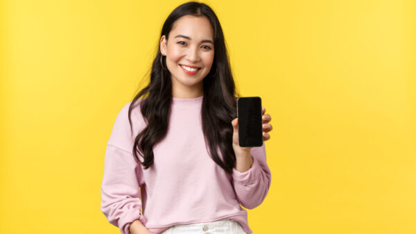 mulher de cabelo preto um pouco acima da cintura e segurando um celular com a tela virada para frente em fundo amarelo