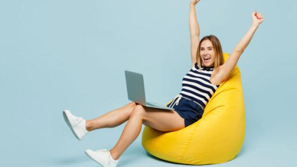 Foto de uma mulher de corpo inteiro usando uma camisa regata listrada casual, sentada em um pufe, com um notebook no colo, fazendo um gesto de vencedora, isolada em um fundo azul pastel simples. Conceito de estilo de vida.