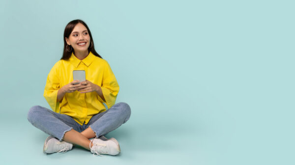 à esquerda está uma jovem sentada no chão de pernas cruzadas e segurando um celular nas mãos em fundo azul