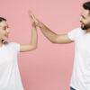 Casal (um homem e uma mulher) alegre, usando camisetas brancas básicas, dando um "high five", isolado em um fundo rosa pastel.