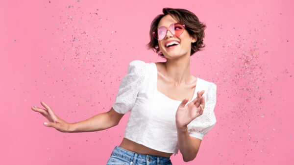 mulher de cabelo curto e óculos de coração feliz e com glitter prateado aos lados em fundo rosa