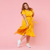 Retrato de corpo inteiro de uma mulher sorridente, ruiva, usando um vestido amarelo, posando, olhando para a câmera, isolada em um estúdio de fundo de cor rosa pastel.