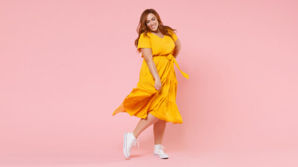 Retrato de corpo inteiro de uma mulher sorridente, ruiva, usando um vestido amarelo, posando, olhando para a câmera, isolada em um estúdio de fundo de cor rosa pastel.