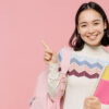 mulher de traços asiáticos carregando um caderno no braço esquerdo e uma mochila rosa clara pendurado no braço direito em fundo rosa