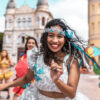 ao centro em destaque tem uma mulher negra com roupas de carnaval e ao seu lado tem mais pessoas fantasiadas para o carnaval em desfoque em um fundo de centro da cidade