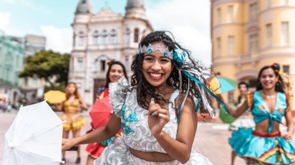 ao centro em destaque tem uma mulher negra com roupas de carnaval e ao seu lado tem mais pessoas fantasiadas para o carnaval em desfoque em um fundo de centro da cidade