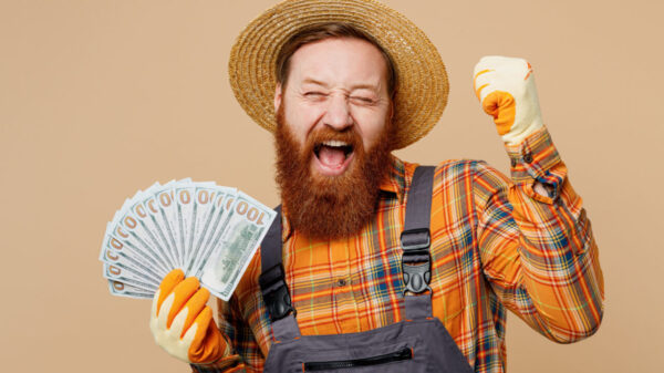 Homem barbudo usando um macacão e um chapéu de palha, segurando notas de dinheiro, fazendo gesto de vencedor, isolado em um estúdio de fundo de cor bege claro liso.