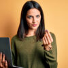 Mulher morena de olhos azuis usando um computador portátil sobre um fundo laranja, fazendo gesto de dinheiro com a mão.